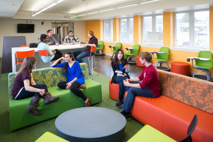 Students relax and chat in a brightly lit common area with colorful furniture, including green, orange, and patterned seating.